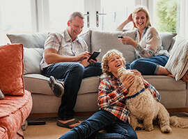 A happy family is sitting in the living room. Their son is sitting on the floor with the family dog licking his face.