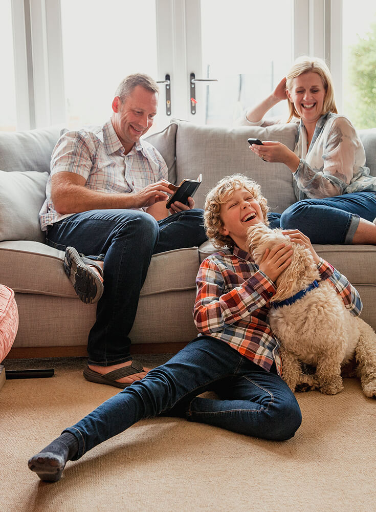 A happy family is sitting in the living room. Their son is sitting on the floor with the family dog licking his face.