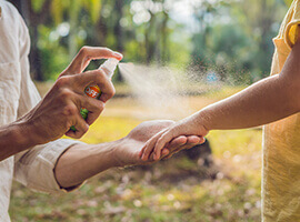 A mom spraying insect repellent on a child's arm outside.