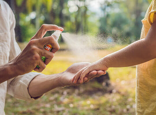 A mom spraying insect repellent on a child's arm outside.